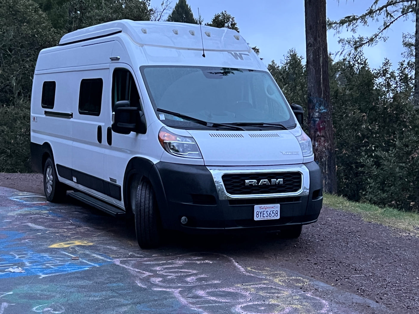 White Winnebago Ram ProMaster camper van, possibly an RV rental, parked on a colorful chalk-covered road.
