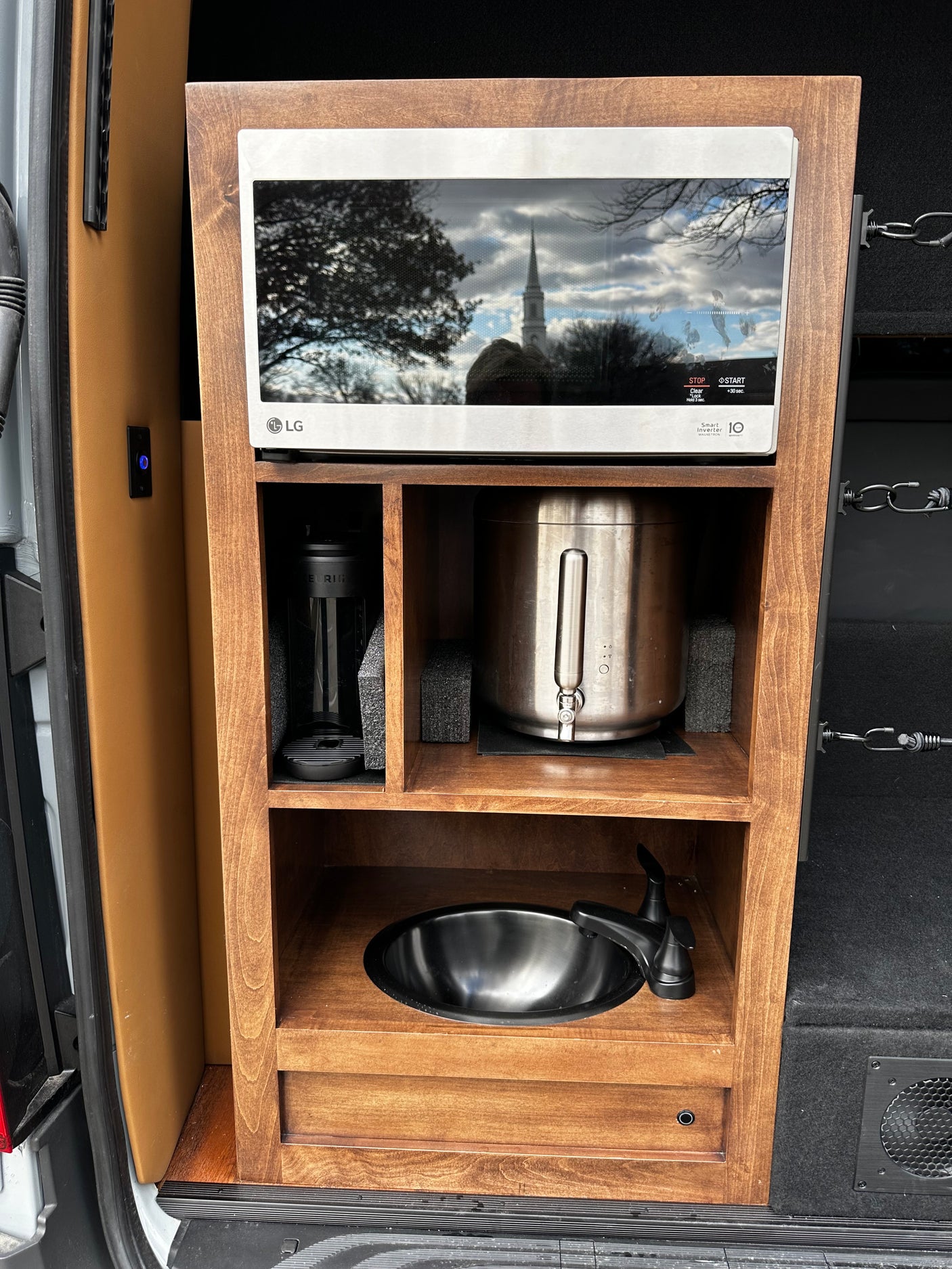 Compact RV rental kitchen setup with an LG microwave, coffee maker, water dispenser, and sink in a custom wooden cabinet.