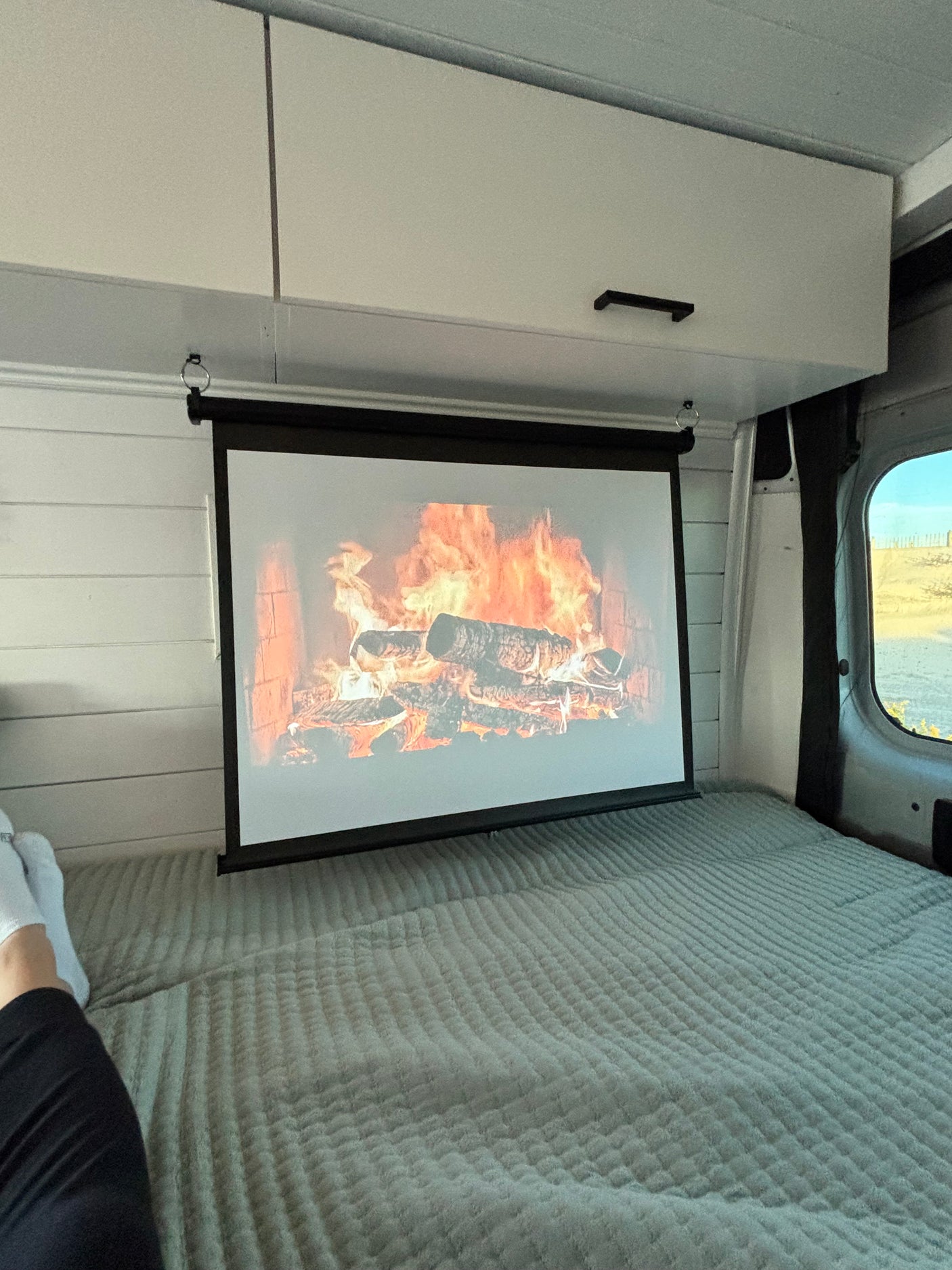 Cozy rv rental interior: fireplace projected on a screen above a bed. A person relaxes with feet up, enjoying the van life ambiance.