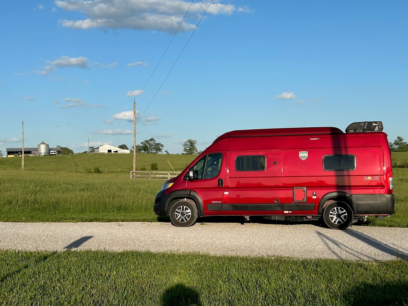 Red Solis camper van for rv rental, parked on a gravel road alongside green fields with distant farm buildings under a blue sky.