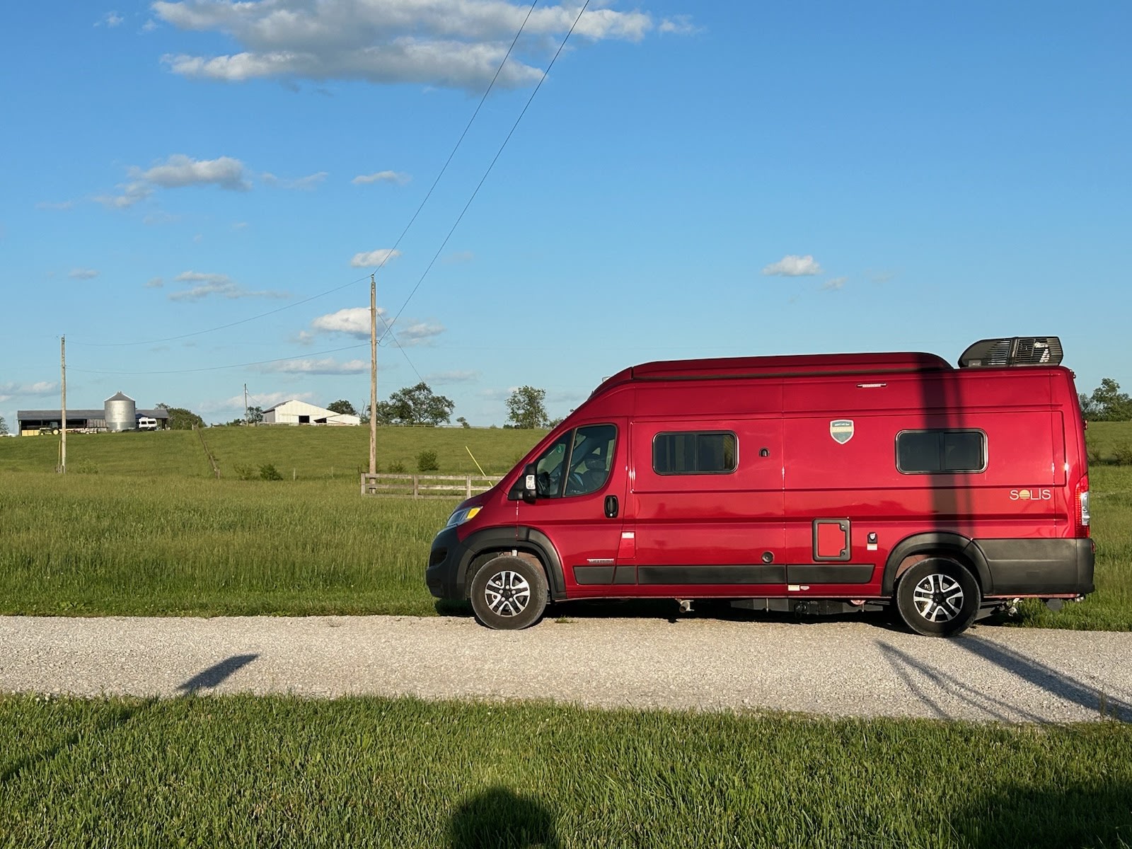 Red Solis camper van for rv rental, parked on a gravel road alongside green fields with distant farm buildings under a blue sky.