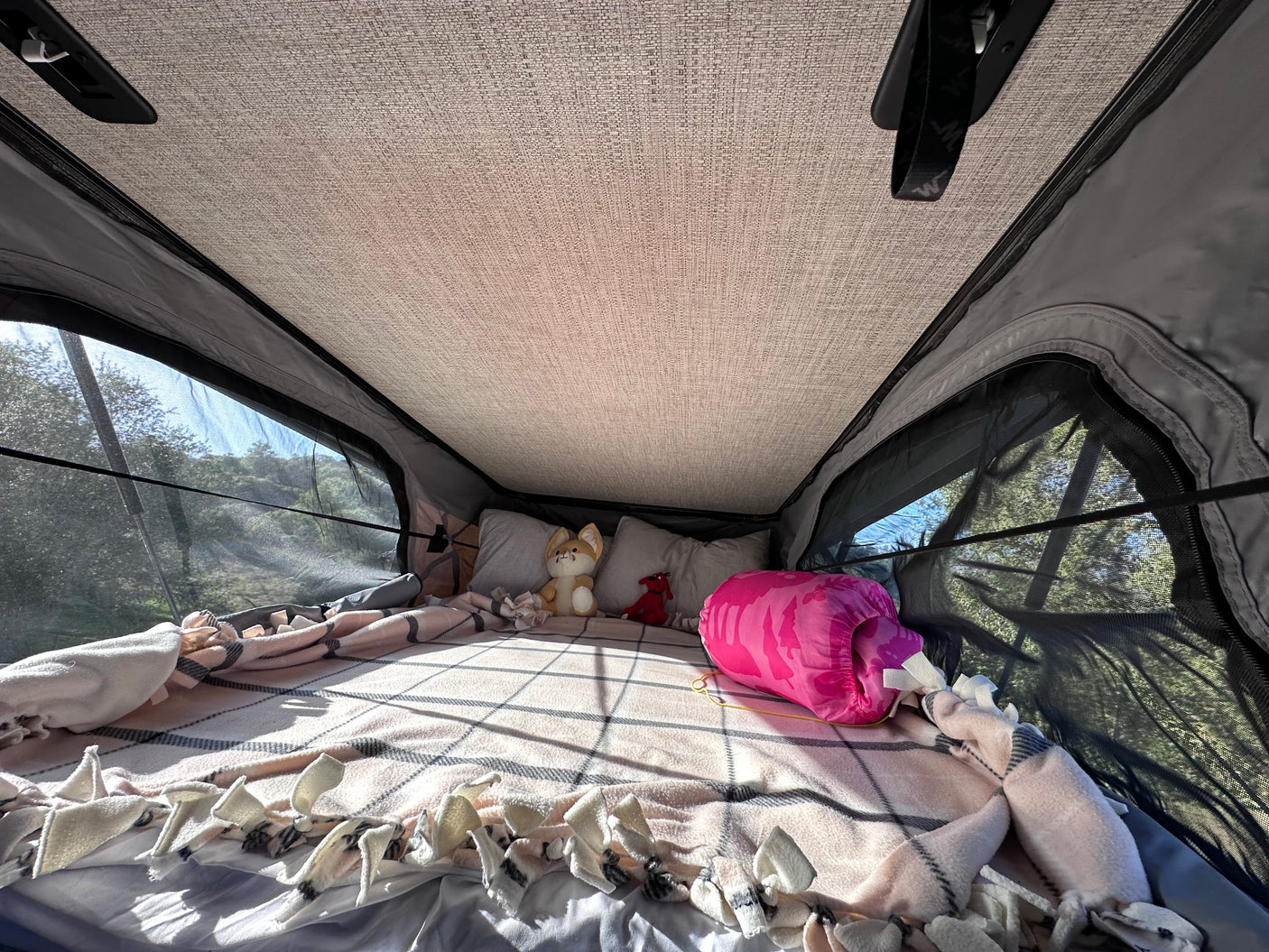 Cozy RV rental interior: bed with blankets, pillows, and stuffed animals, with mesh windows offering views of lush trees.