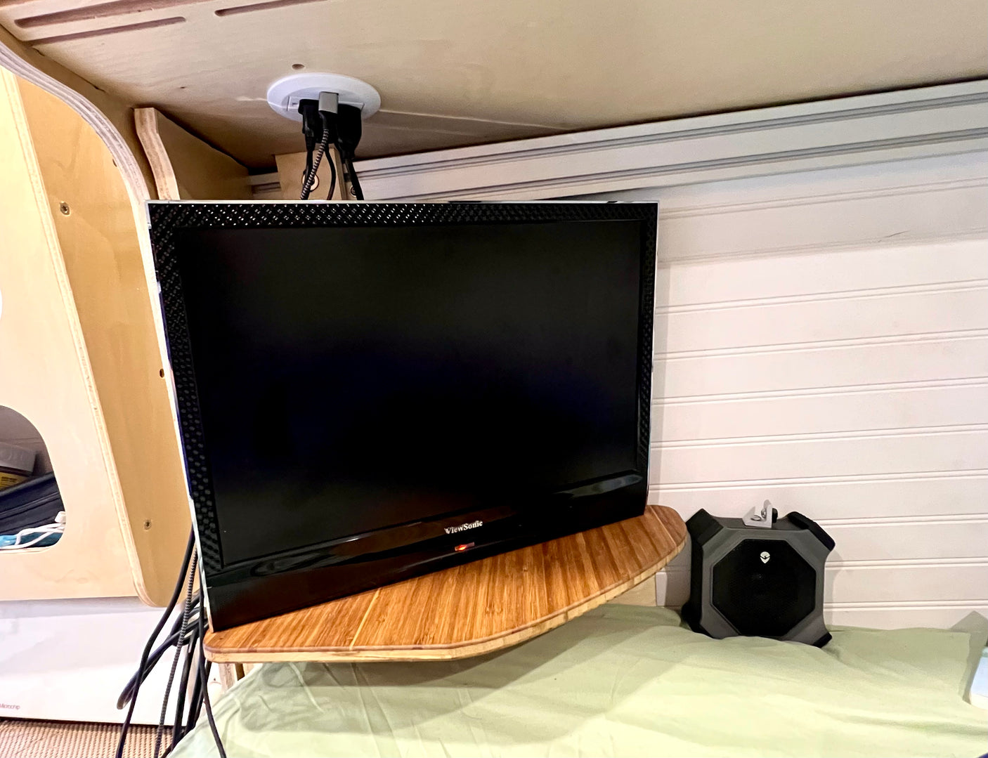 An RV rental's cozy entertainment corner: ViewSonic TV on a wood shelf, cables discreetly routed, beside an Alienware speaker on light green bedding.