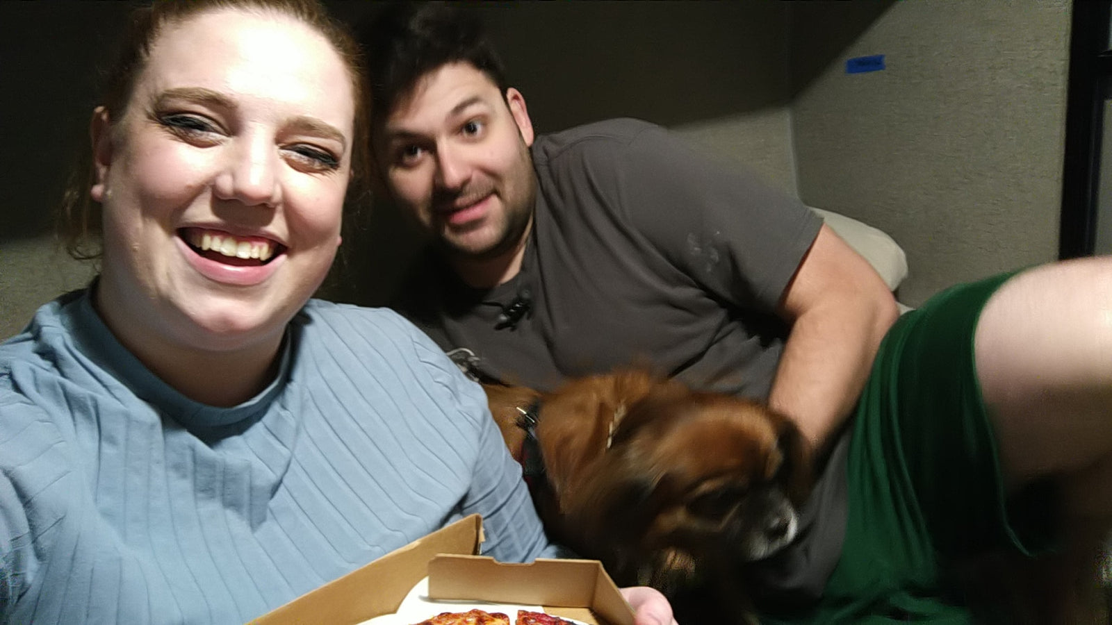 Cozy night in an RV rental for a smiling couple and their dog, enjoying pizza together.