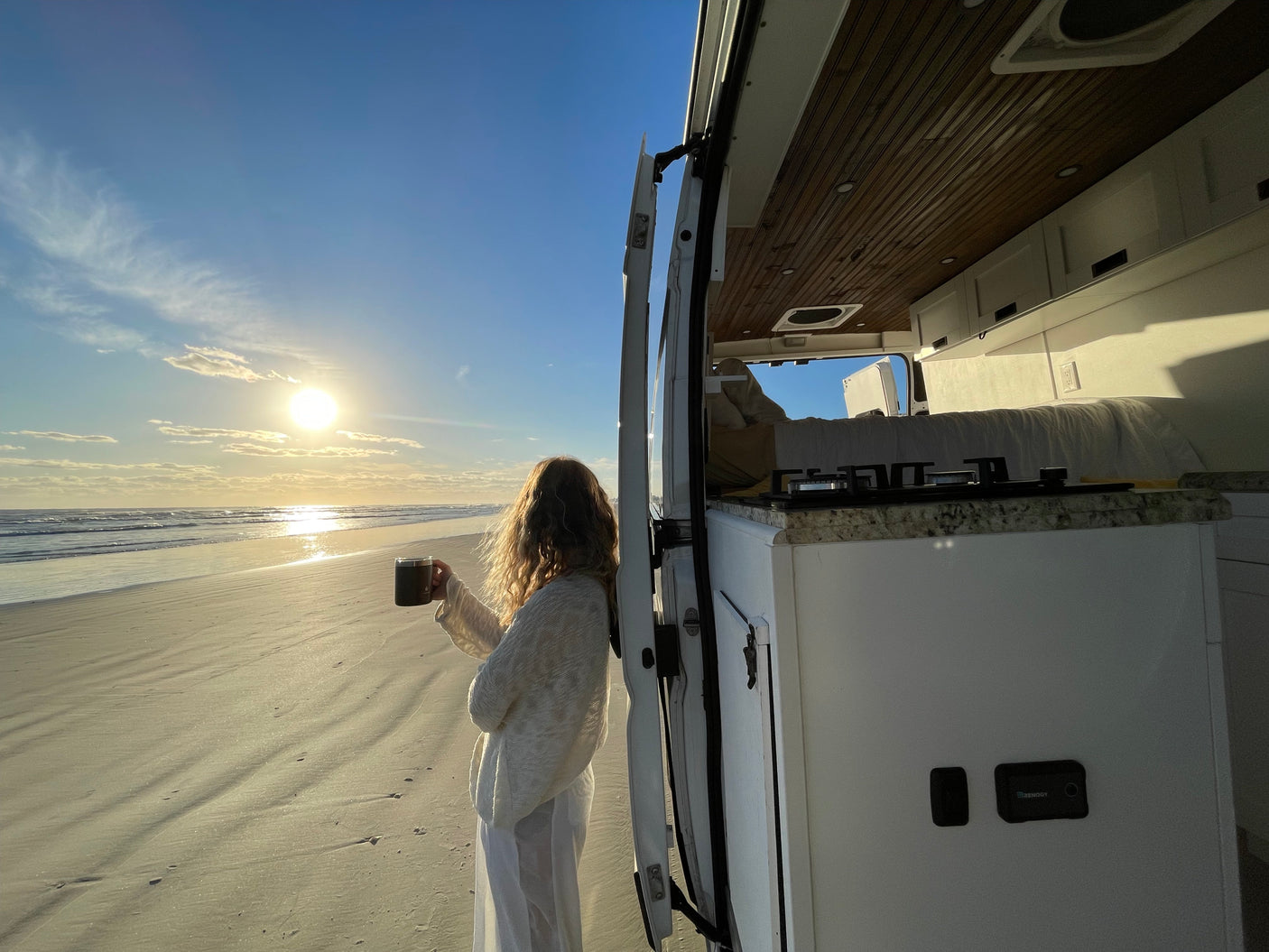 Woman sips coffee, watching a beach sunrise from her RV rental. Campervan open to sandy shore.