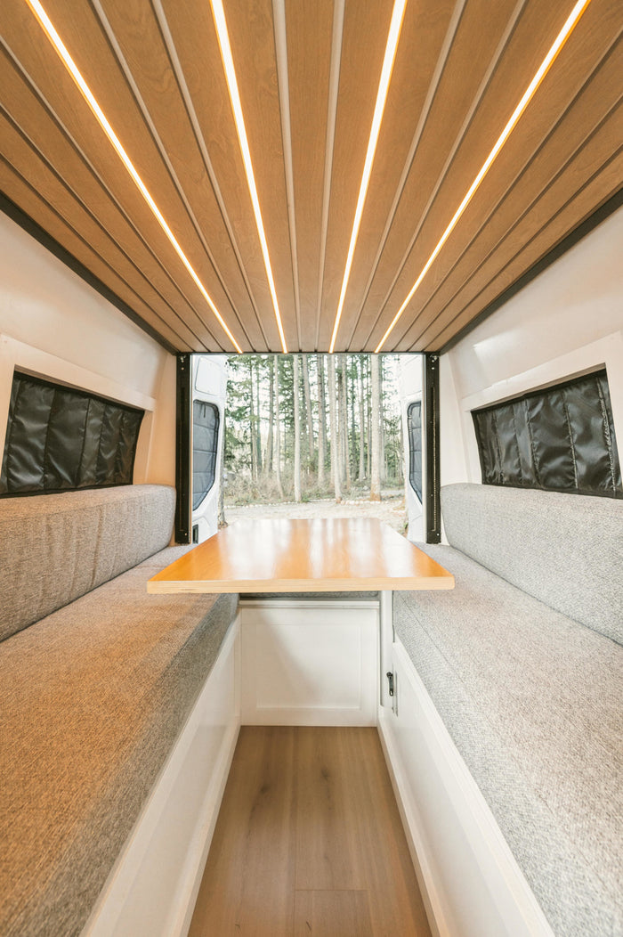 RV rental interior features: wood ceiling with linear LEDs, comfy grey benches, and a dining table. Rear doors reveal a serene forest view.