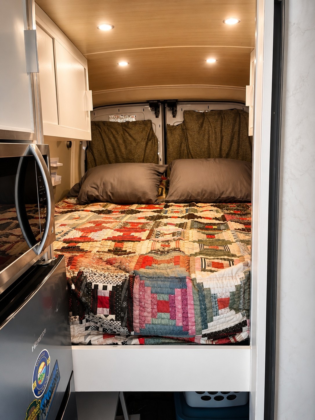 Inside a cozy RV rental: a colorful patchwork bed, wood ceiling, and compact kitchen with white cabinets and a microwave.