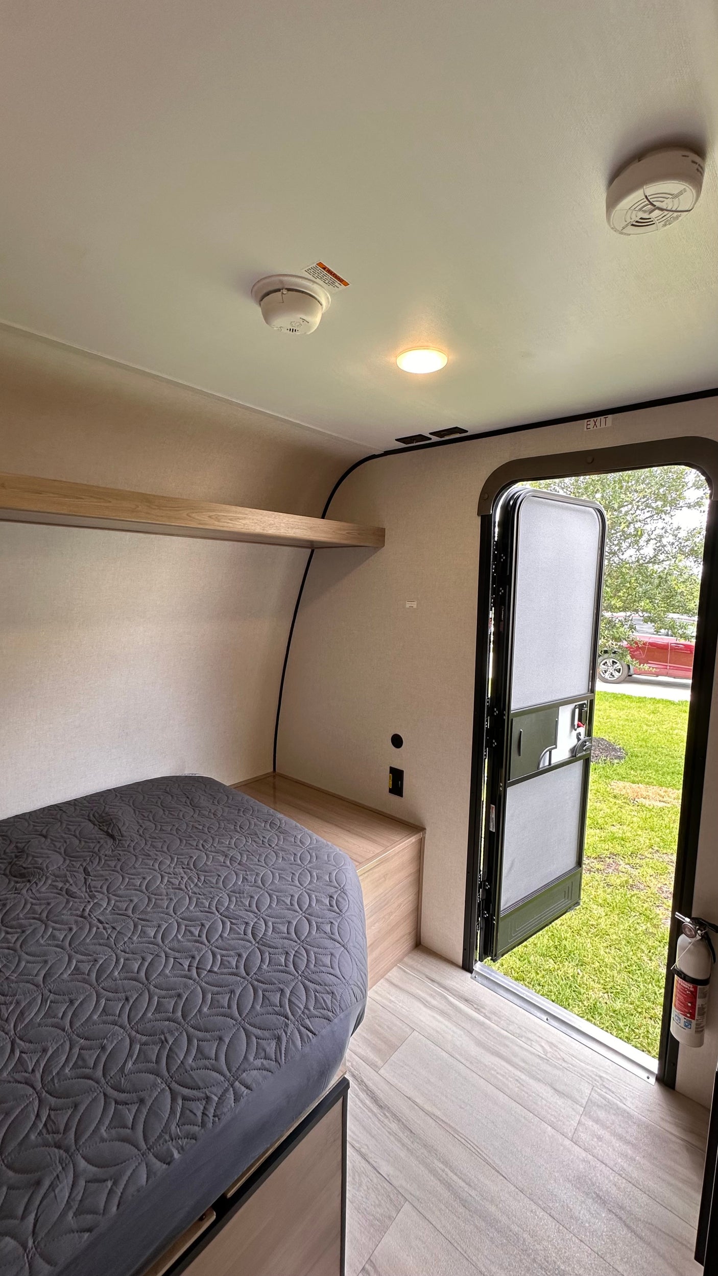 RV rental interior: cozy bed, wooden shelf, smoke alarms, fire extinguisher by the open door to the outdoors.