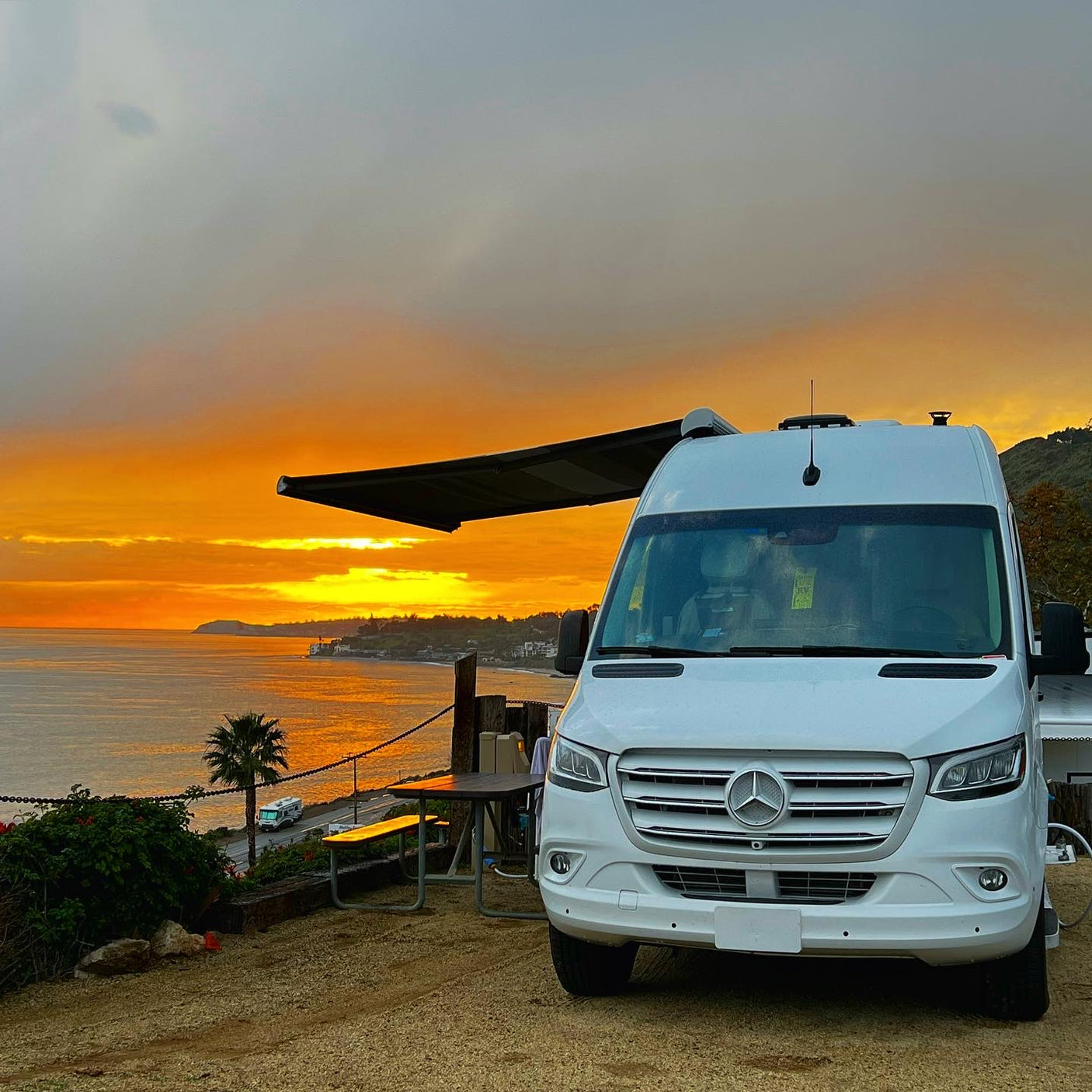 White RV rental (Sprinter camper van) parked on a bluff, showcasing a vibrant sunset over the ocean. Awning out, with a picnic area.