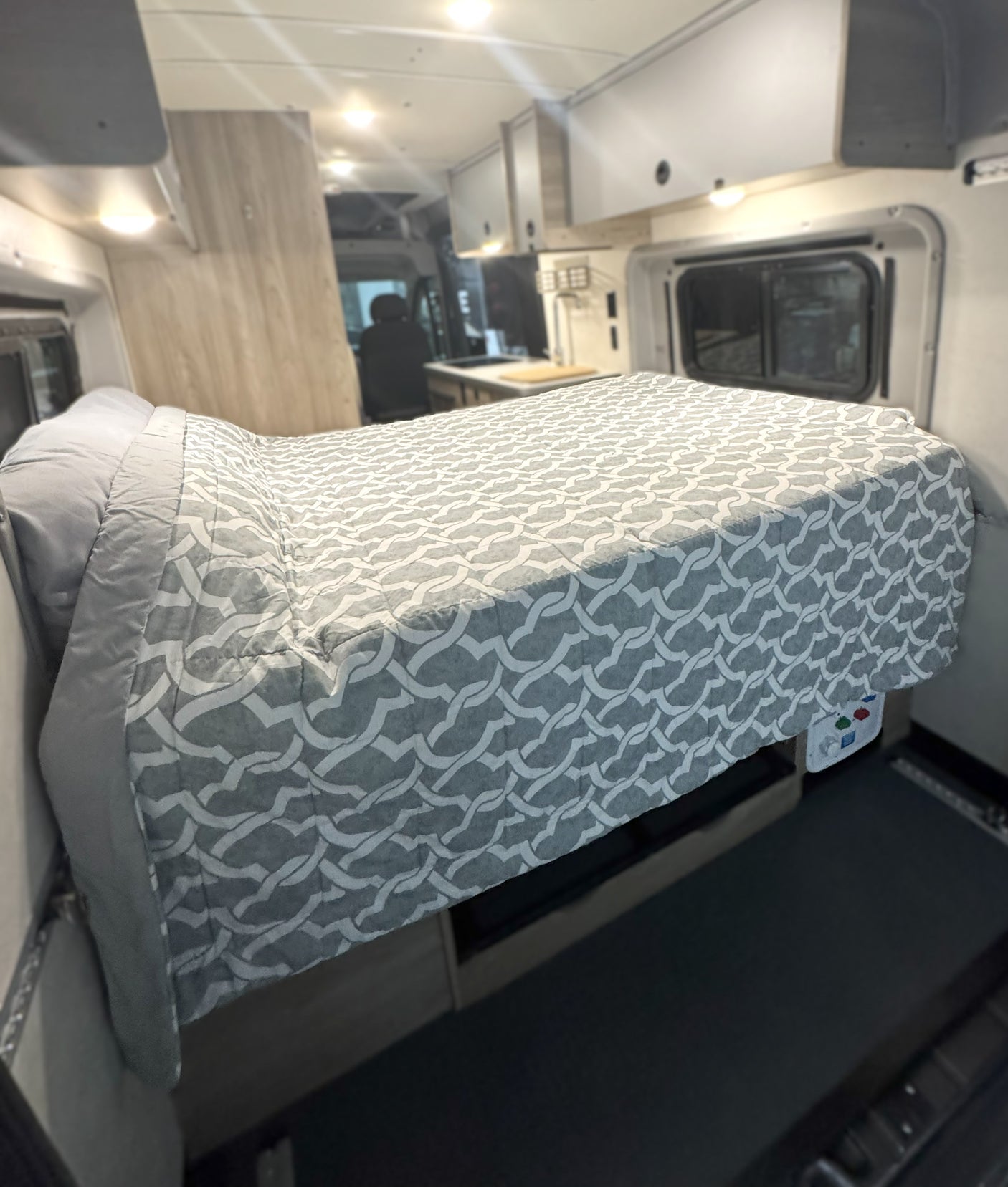 RV rental interior featuring a comfy bed with a gray patterned duvet, compact kitchen, and large side window for road trips.