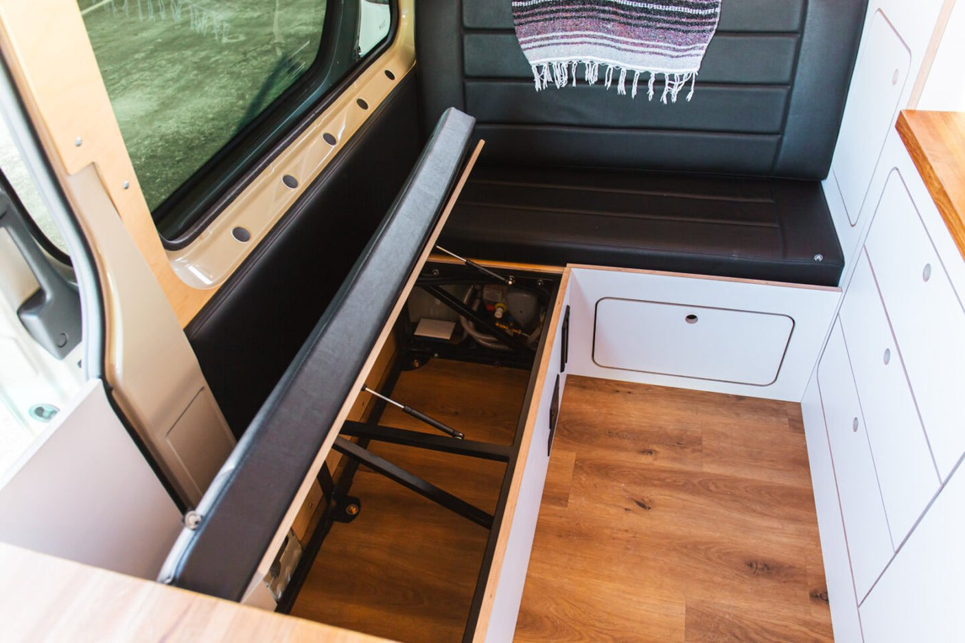 Inside an RV rental: a custom campervan interior with a lift-up bench seat for storage, sleek white cabinets, and wood flooring.