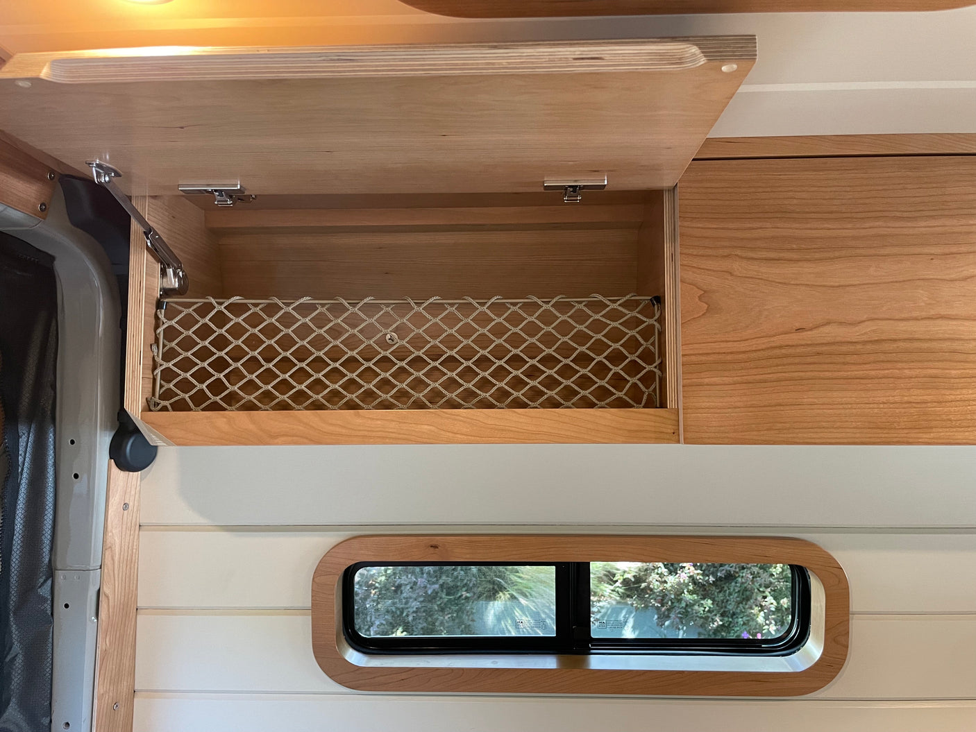 Custom wood interior of an RV rental with an open overhead cabinet, storage net, and small window, ready for travel.