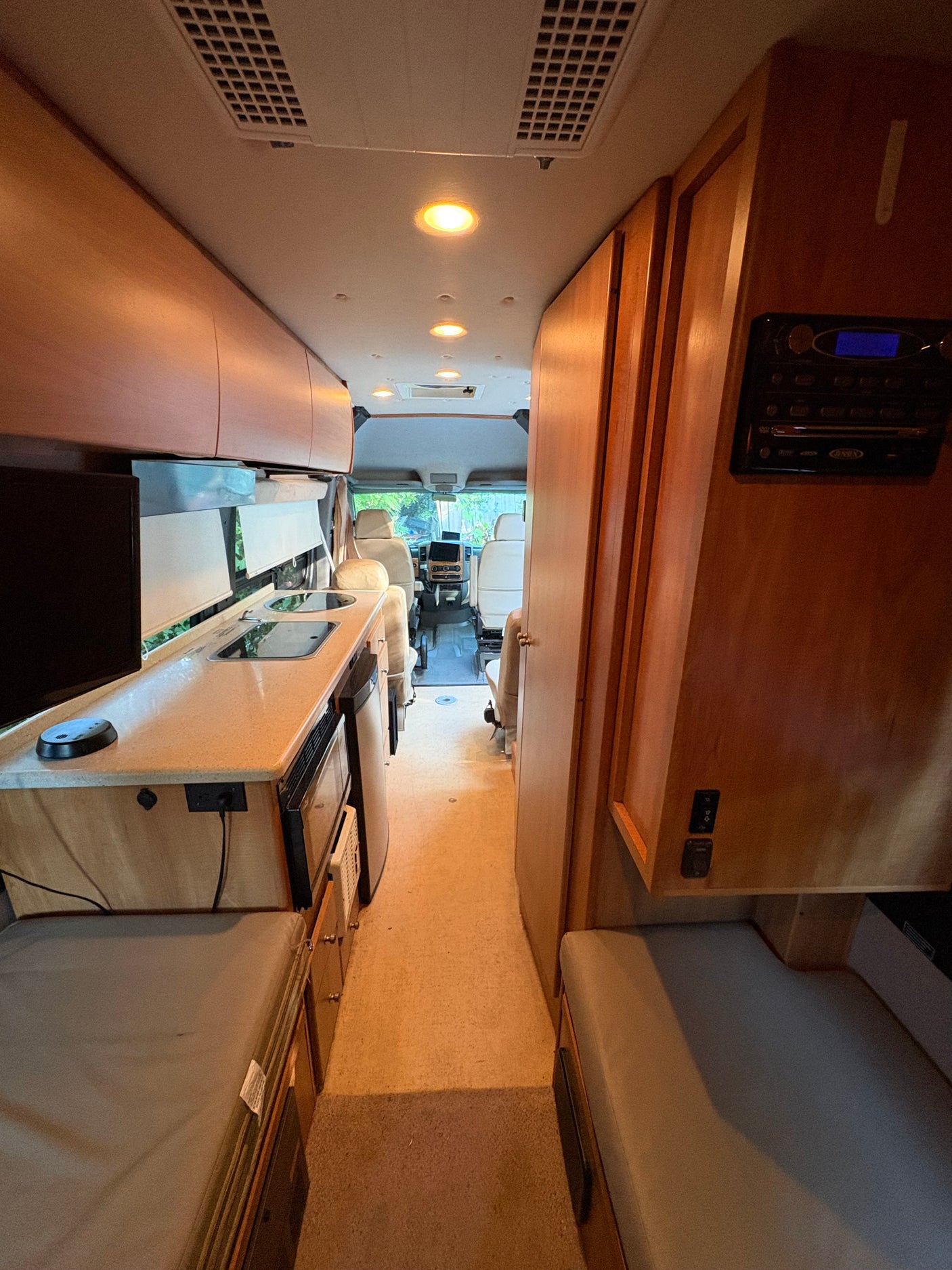 RV rental interior: A compact kitchen with sink and stovetop, overhead storage, seating, and aisle leading to the cockpit.