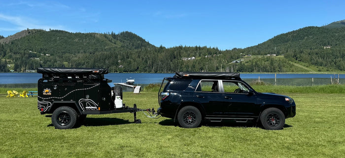 Black SUV & rugged overland trailer parked lakeside. Ideal RV rental for off-grid adventure.