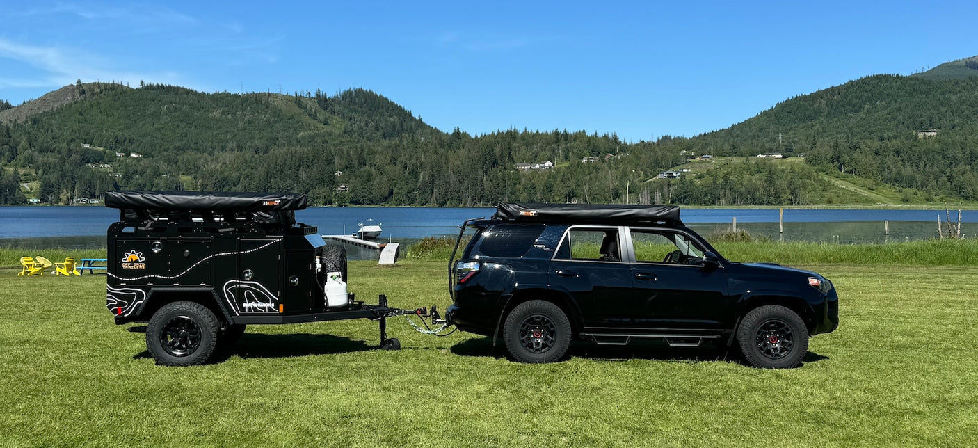 Black SUV & rugged overland trailer parked lakeside. Ideal RV rental for off-grid adventure.
