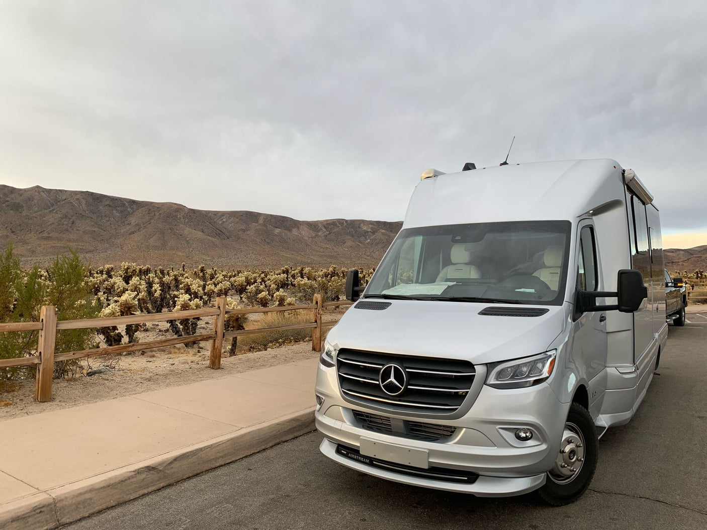 An Airstream rv rental parked roadside near a Cholla Cactus Garden with desert mountains under a cloudy sky.