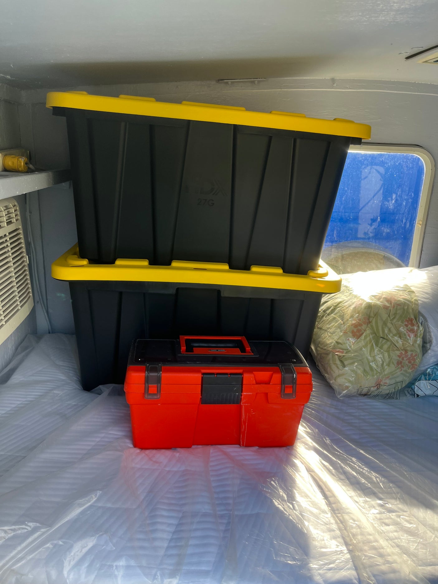 RV rental: Stacked black-yellow storage bins & an orange toolbox are neatly placed on a plastic-covered mattress. Bedding visible.