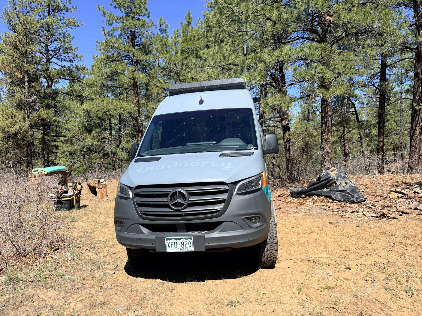 Outdoorsy rv rental Mercedes Sprinter camper van parked on a dirt road in a sunny pine forest with camping gear.