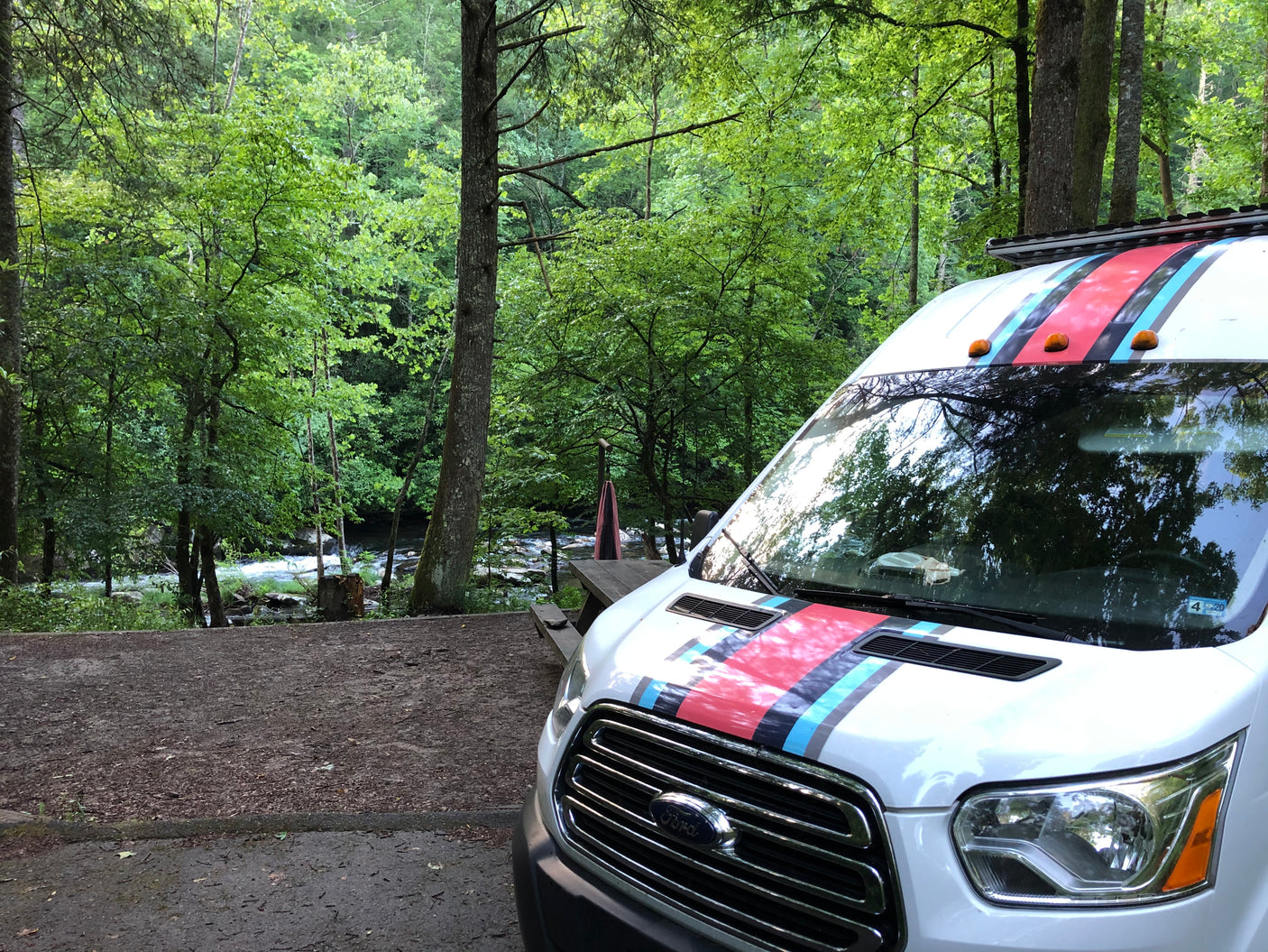 White Ford Transit rv rental, styled with stripes, at a scenic riverside campsite surrounded by lush green trees.