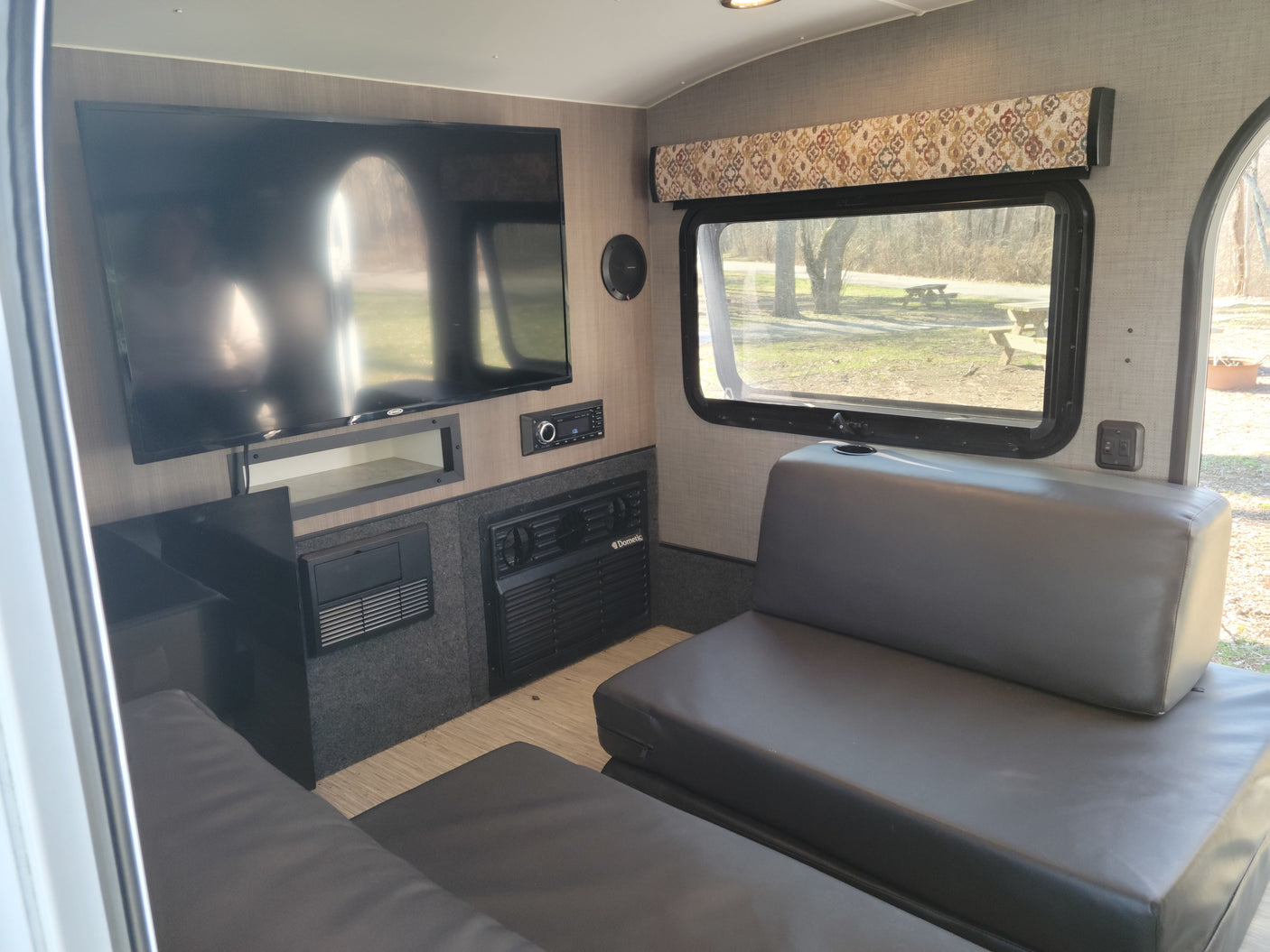 Modern **RV rental** living space: TV, stereo, Dometic AC/heater, and leatherette sofa overlooking a campsite.