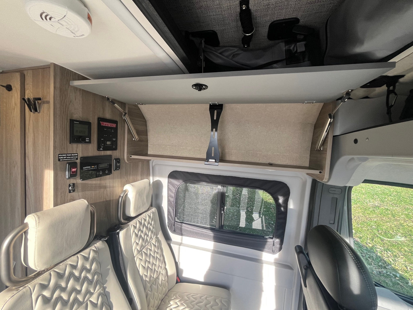 Explore this **rv rental** interior featuring cream quilted seats, a window, open overhead storage, and control panels for comfort and convenience.