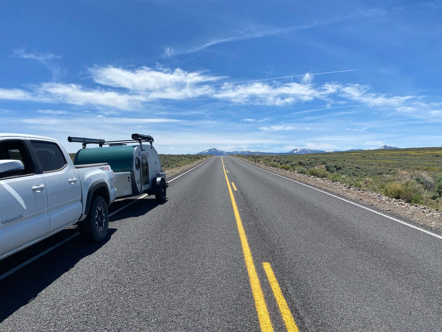 White pickup and attached teardrop camper (an RV rental) on a long, open highway leading to snowy mountains.