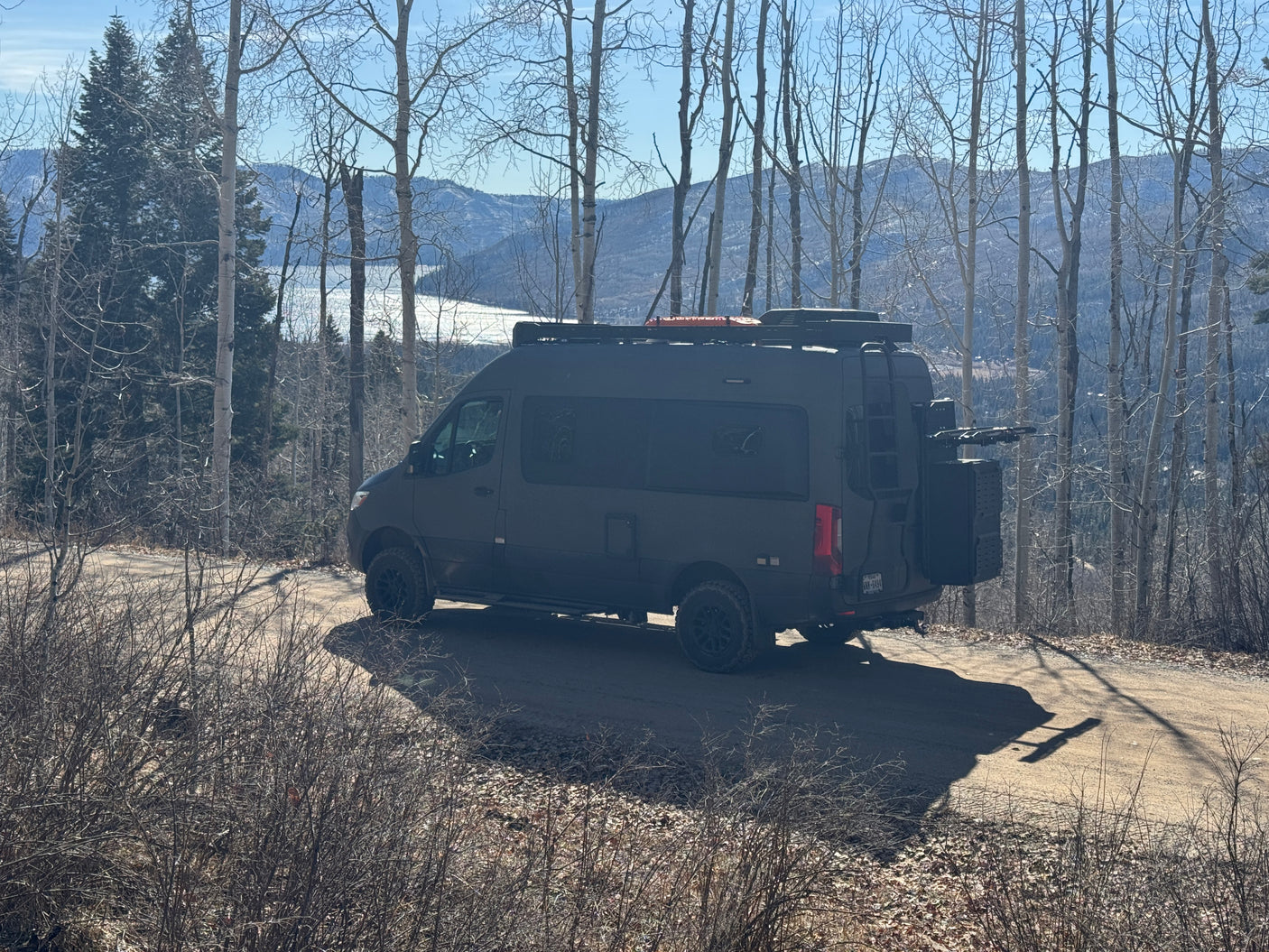 Dark adventure camper van, perfect for an rv rental, parked on a dirt road overlooking a scenic lake and mountains.