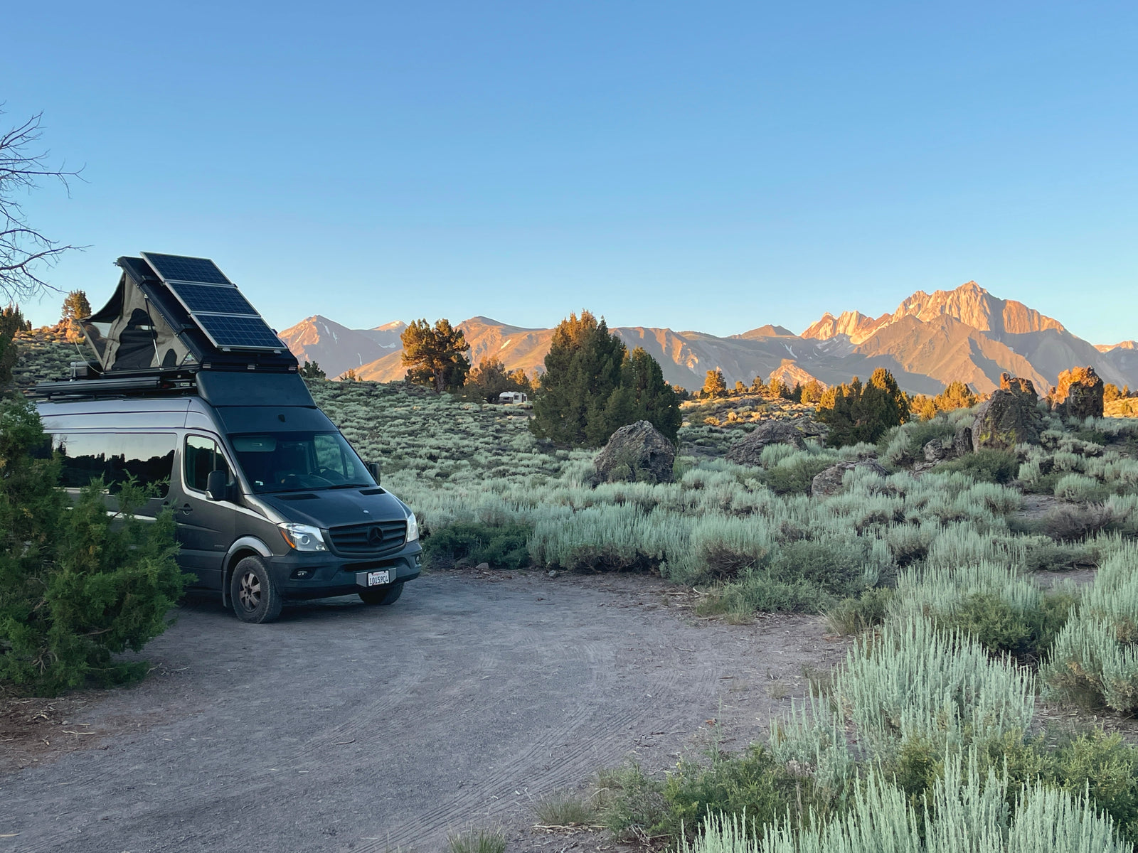 An RV rental camper van with solar panels is parked on a dirt patch amidst sagebrush, with sunlit mountains behind.
