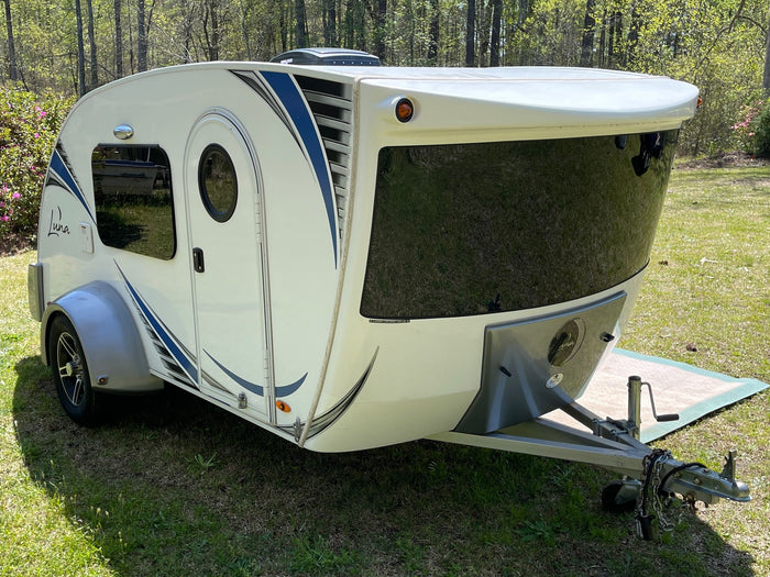 is parked on green grass by a scenic pond.|Modern white teardrop camper