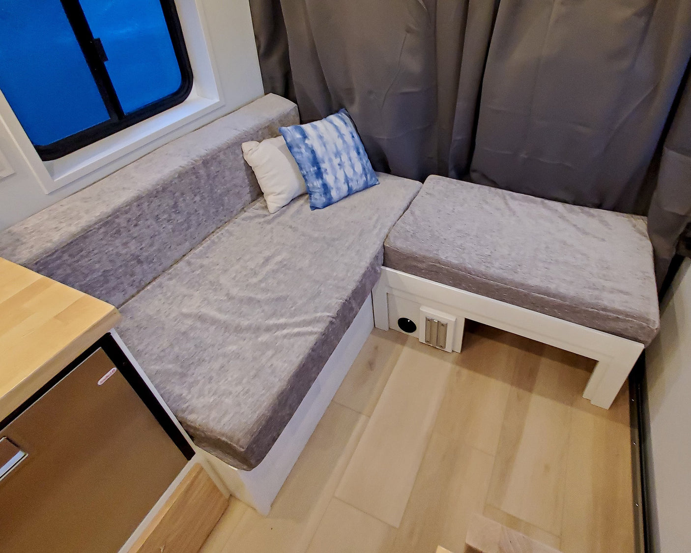 Cozy RV rental interior features L-shaped seating with grey cushions, blue tie-dye and white pillows, and a window.