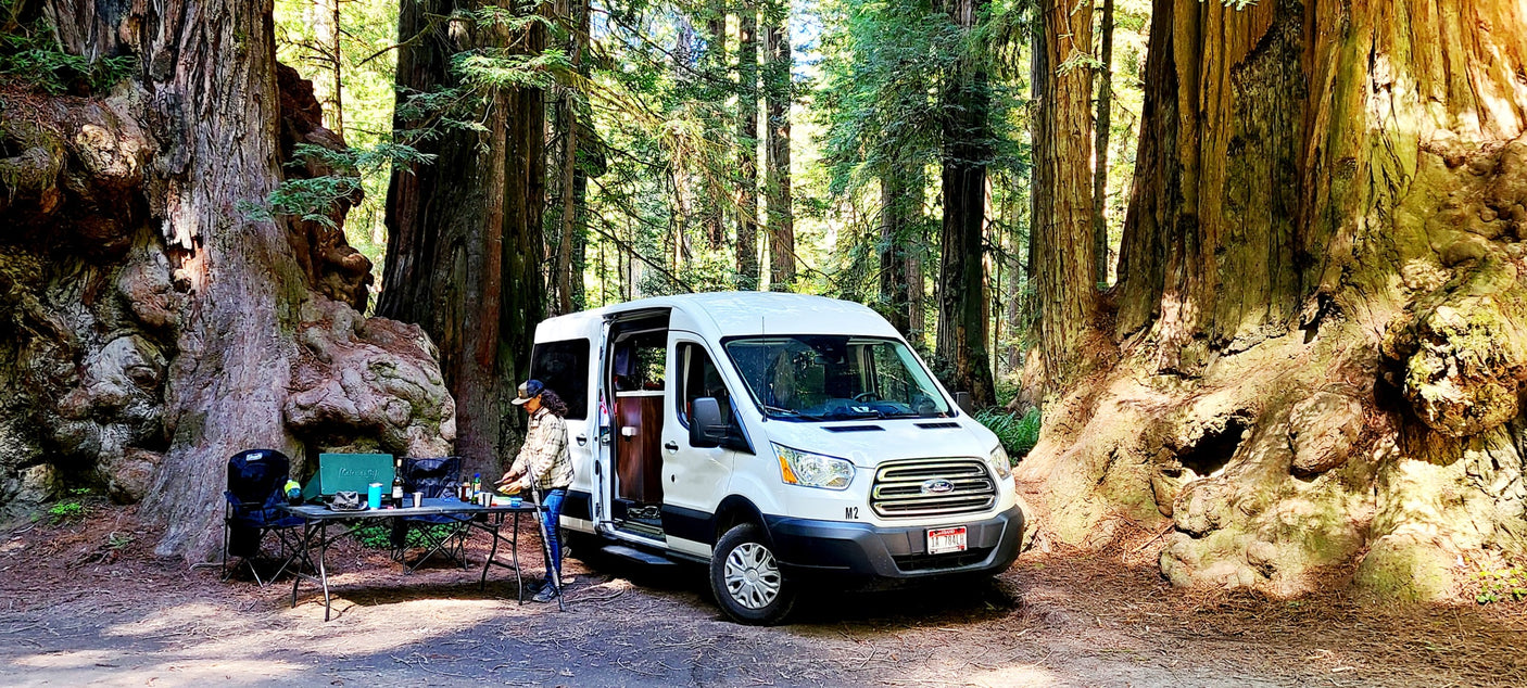 An RV rental van is parked in a lush redwood forest campsite. A person prepares food at an outdoor table amidst giant trees.|Inside an RV rental