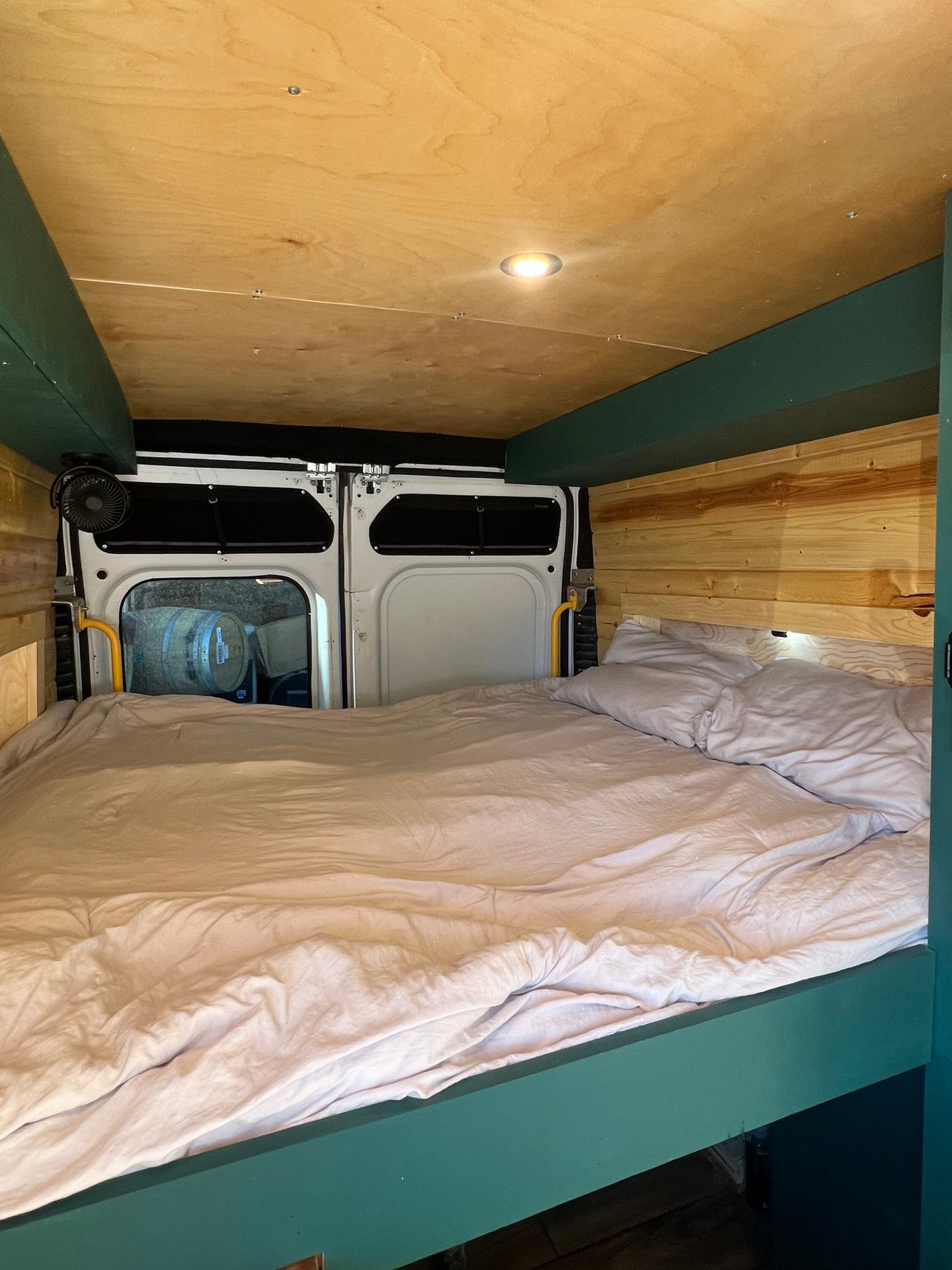 Inside a cozy camper van rv rental with a made bed, wooden walls & ceiling, and rear doors opening to barrels outside.