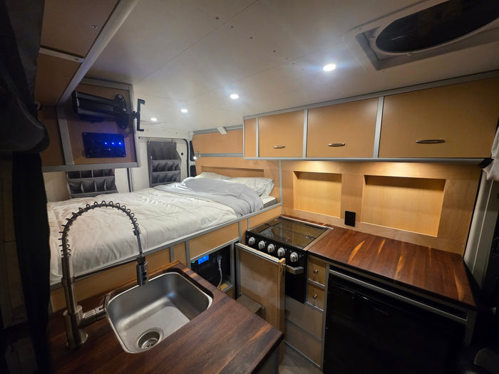 This RV rental interior shows a compact bed, full kitchen with sink/stove, and overhead cabinets for comfortable travel.
