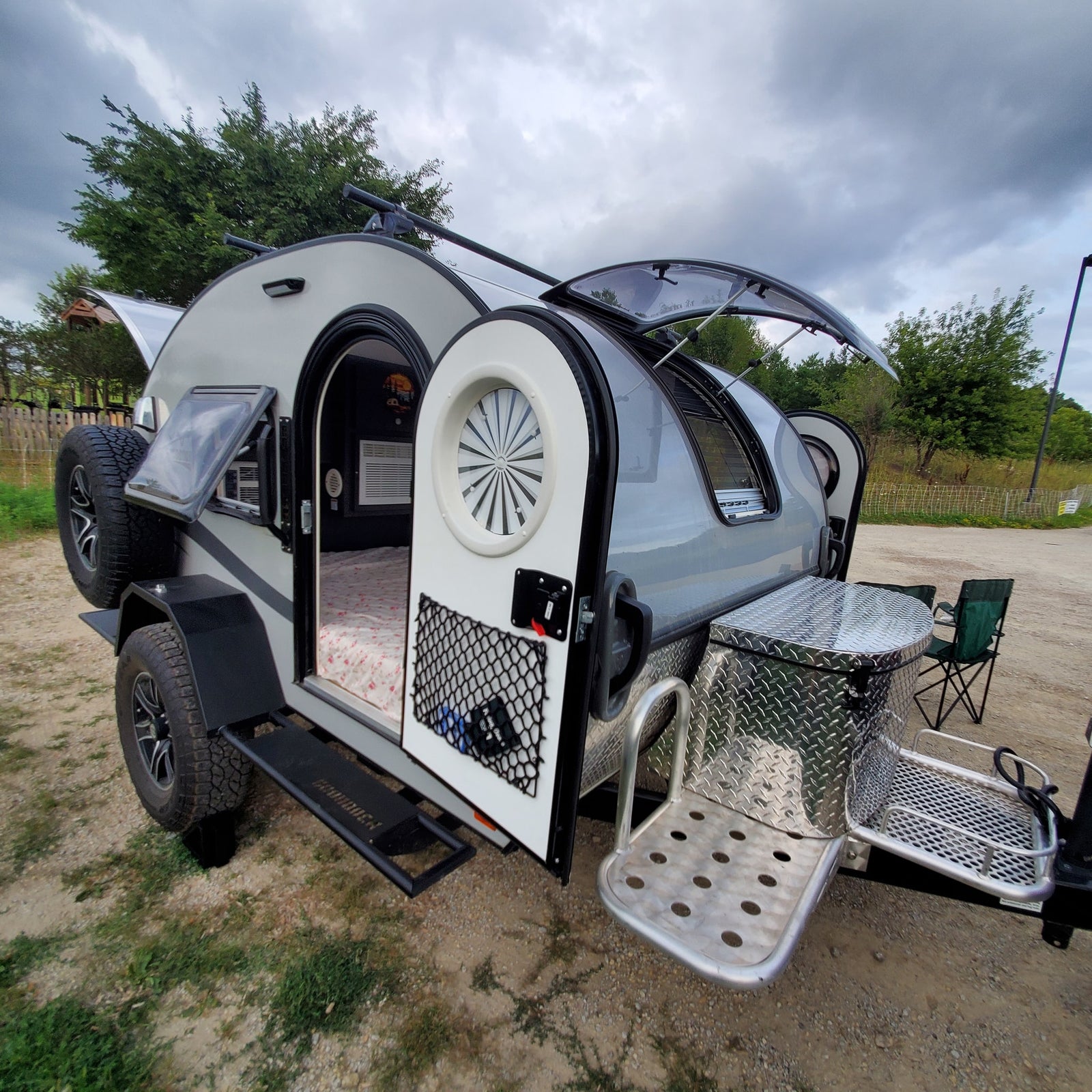 Compact RV rental: a modern teardrop camper with open door revealing bed, plus outdoor storage and steps.