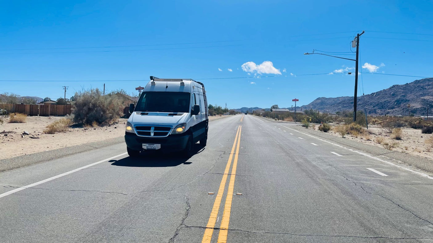 A white/black RV rental van travels a long desert road. Mountains, power lines, and dry brush are visible under clear blue skies.