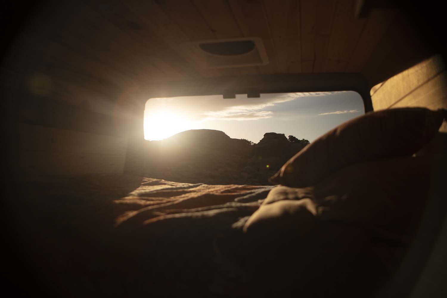 Wake up to desert views from your RV rental! Cozy bed inside, with sun over mountains through the window.