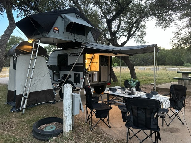 Modern RV rental camping setup: rooftop tent, outdoor kitchen, and dining area under an awning in nature.