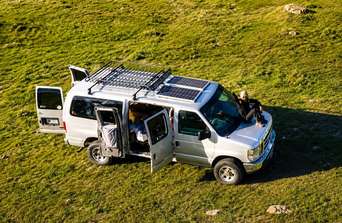 RV rental camper van with solar panels on a grassy hill. Doors open, person on hood. Ideal for outdoor adventures.