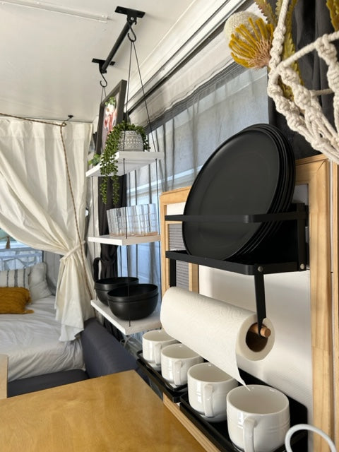 Compact RV rental interior featuring a neat kitchenette with hanging mugs, plate rack, and paper towels, next to a cozy bed.