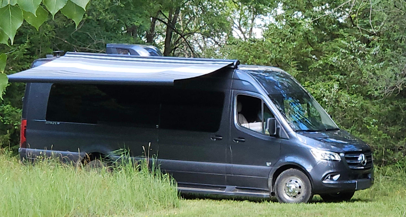 Dark gray Mercedes Sprinter camper van RV rental with extended awning, parked in grass outdoors.