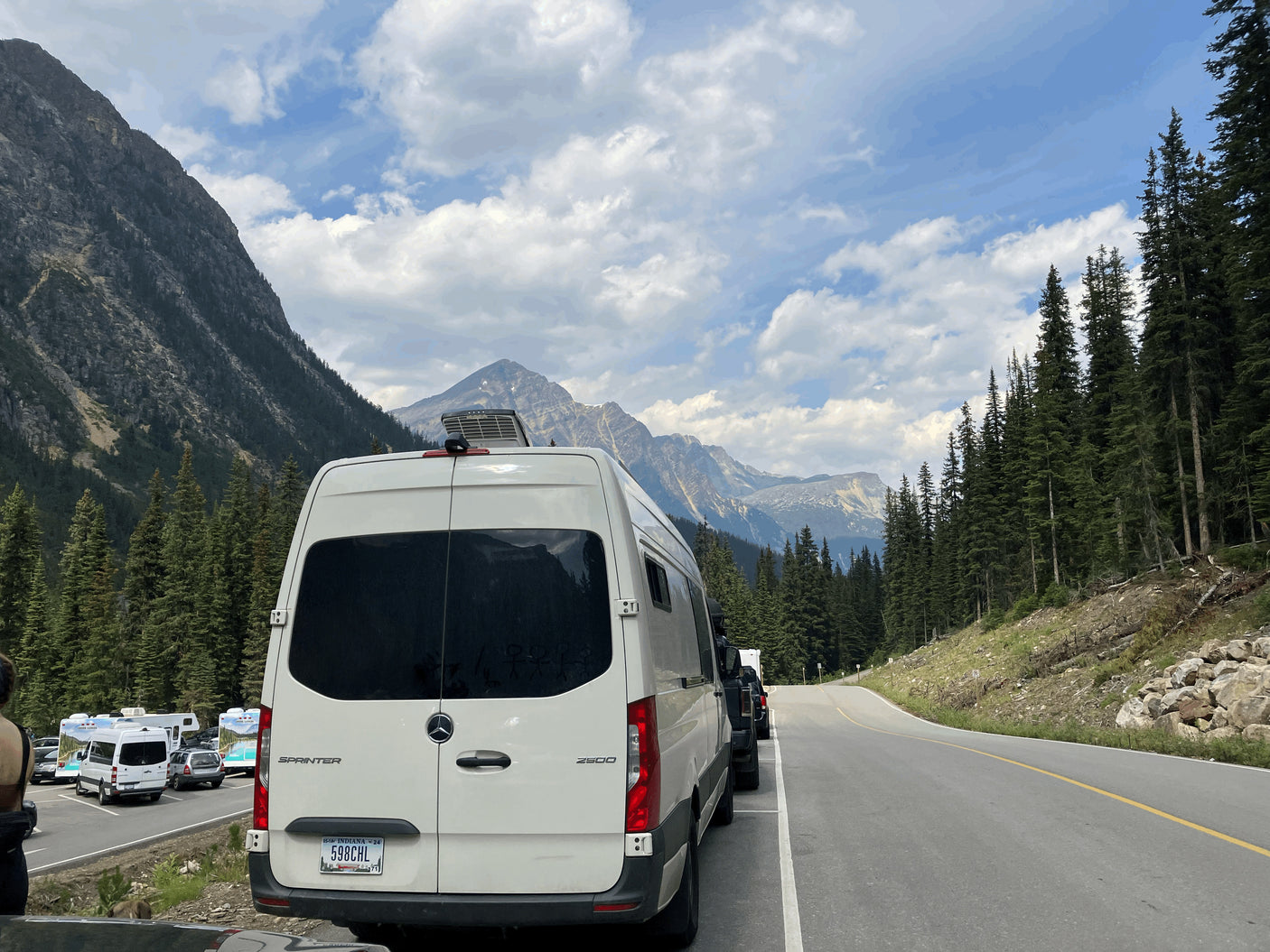 An rv rental van with Indiana plates rests by a mountain road, with other RVs parked nearby in a stunning landscape.