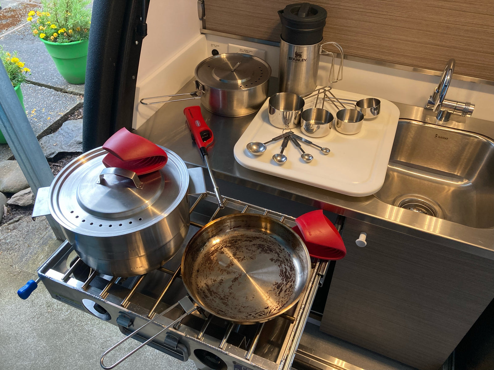 Compact RV rental kitchen with pull-out stovetop, pots, pan, measuring tools, Stanley mug & sink for outdoor cooking.