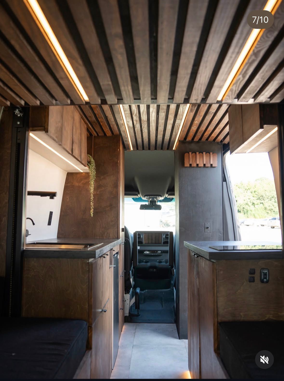 The interior of a stylish RV rental. A slatted wooden ceiling with LED lights illuminates a compact kitchen, seating, and custom cabinetry, leading to the driver's cab.