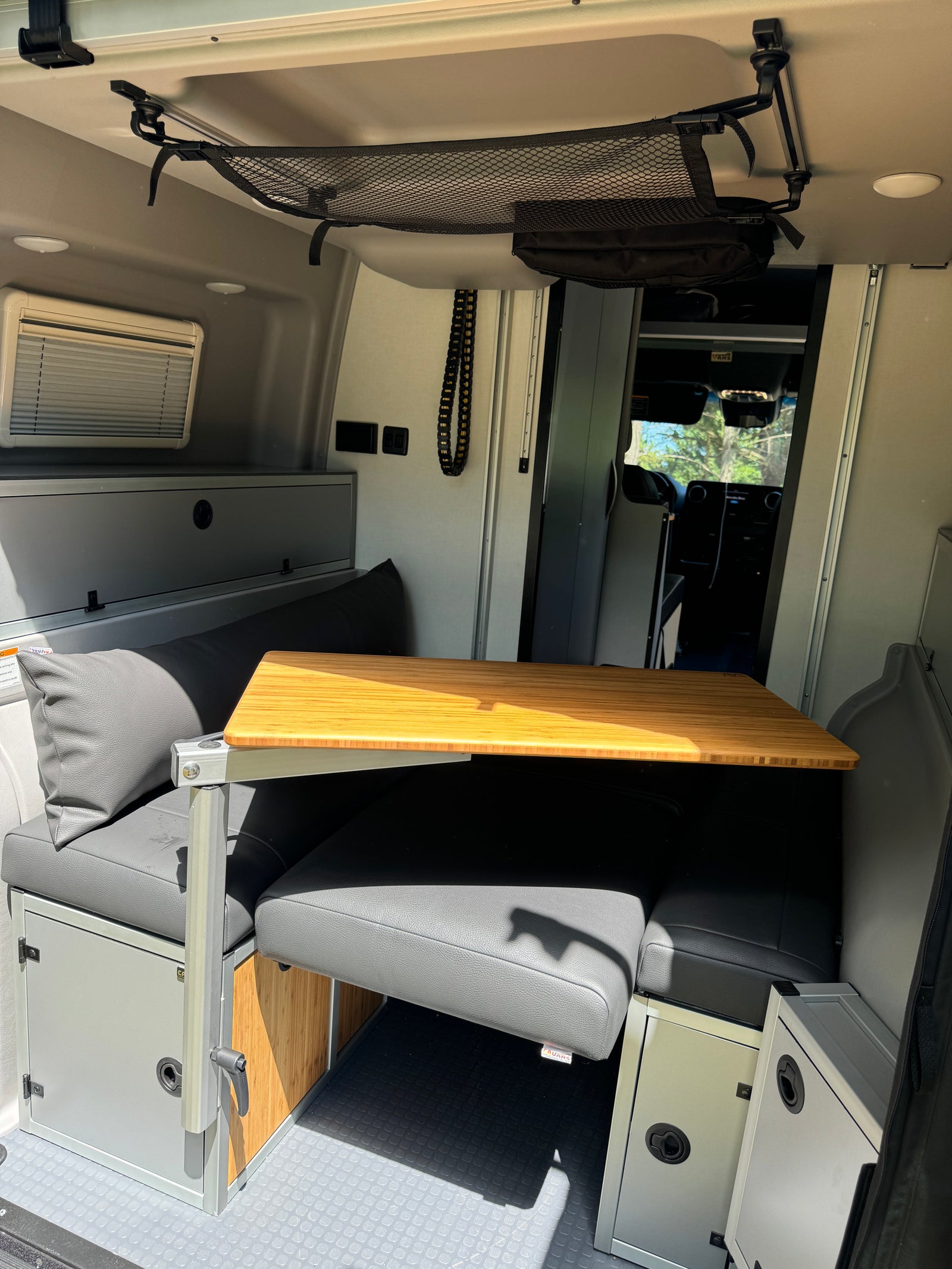 This RV rental features a compact interior dining area with a bamboo table, gray cushioned benches, and overhead storage net.
