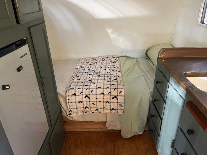 Cozy RV rental interior with a bed, bat-patterned blanket, compact fridge, and a small kitchenette with wooden counters.