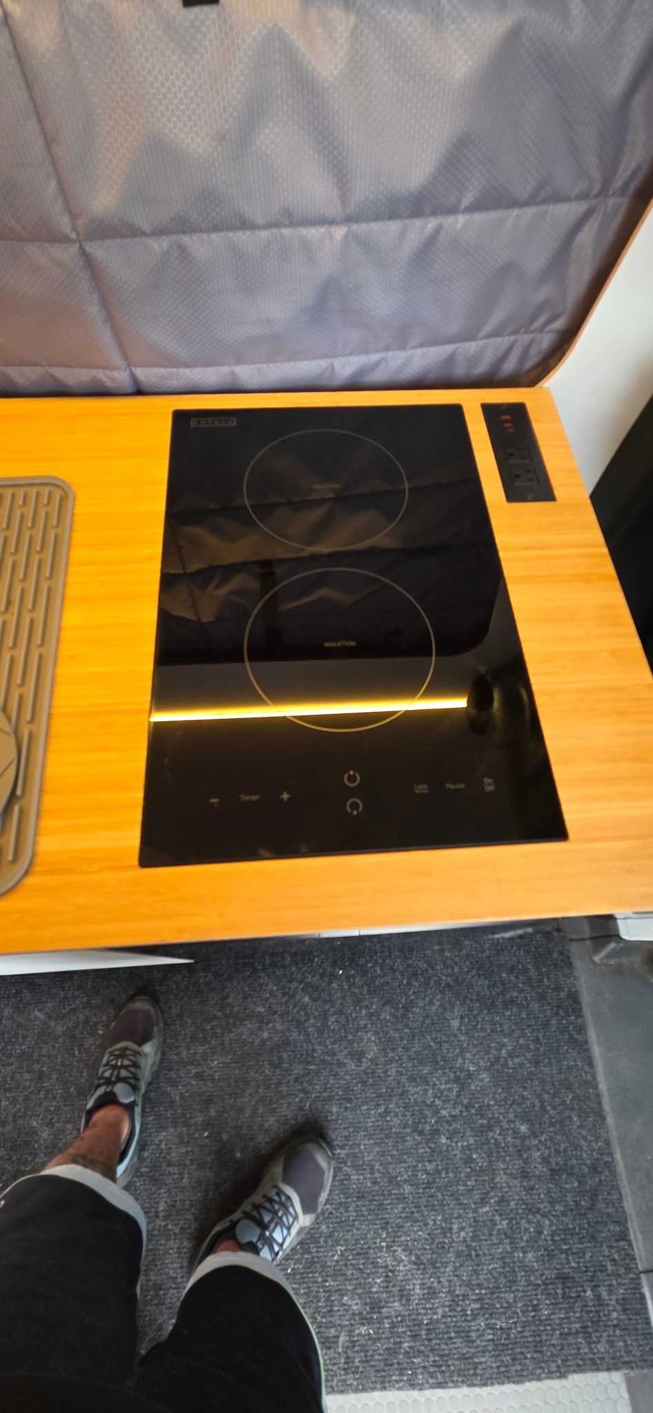 RV rental kitchen: sleek induction cooktop on wood counter with outlets and a drying mat. Person's feet on gray carpet. — photo 11