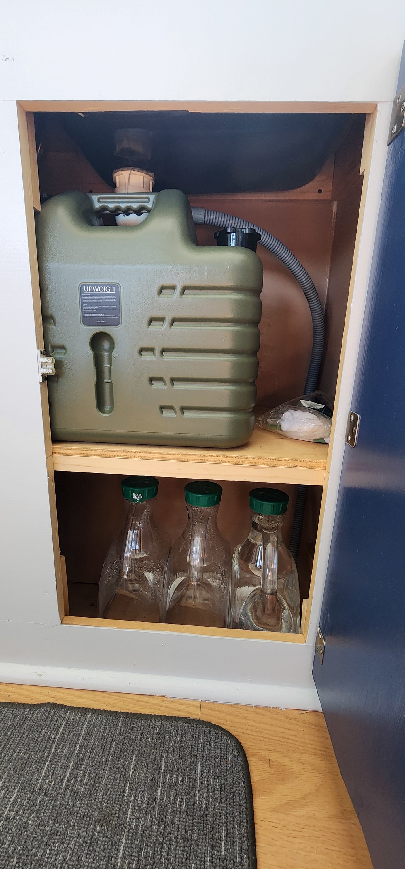 RV rental: Under-sink view showing a large green water tank (UPWOIGH) & glass bottles for compact, off-grid water storage.