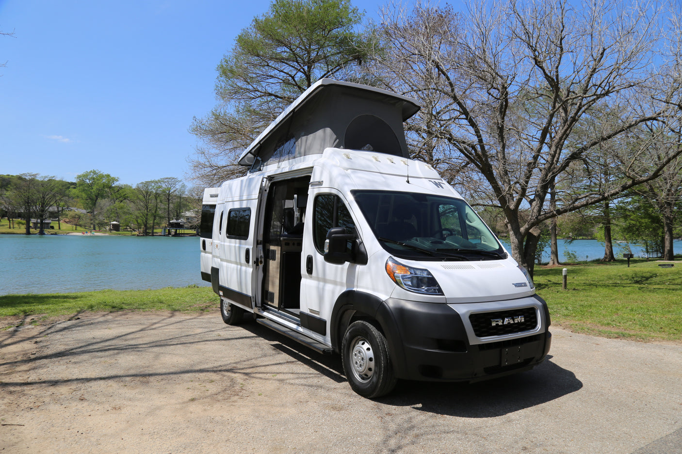 White pop-top RV rental parked lakeside, door open to serene blue water and green trees. Perfect for adventure.