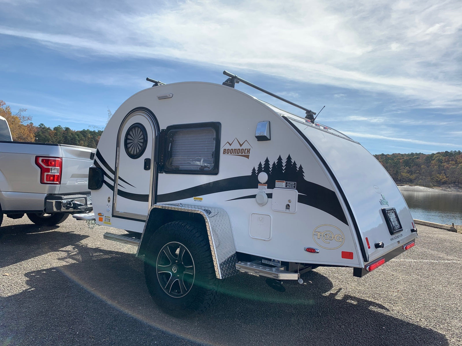 This white Boondock teardrop travel trailer, an ideal RV rental for outdoor adventure, is parked lakeside, ready for camping.