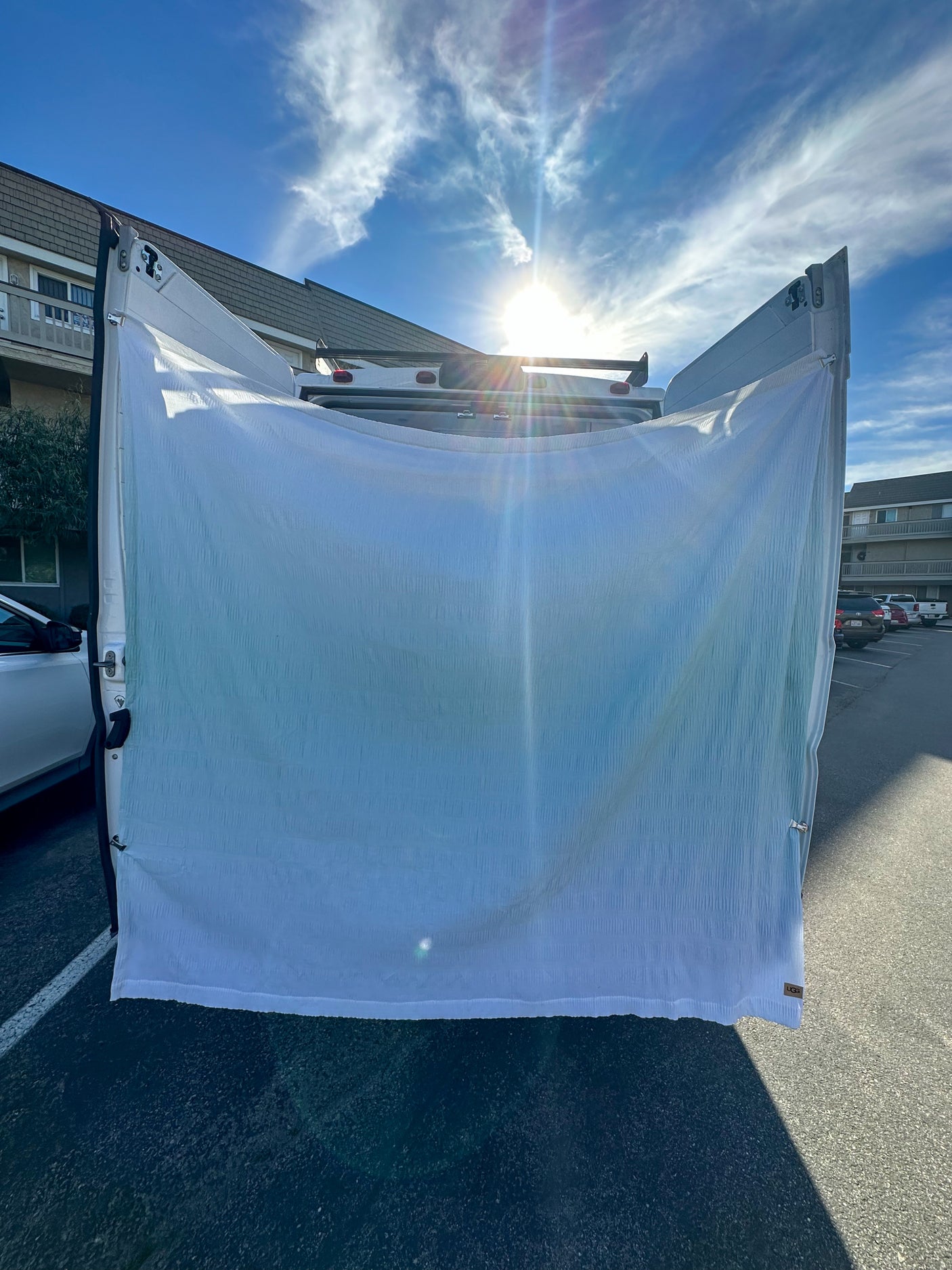 Bright sun shines over an **RV rental** van with a light blue privacy curtain hanging across its open rear doors in a parking lot.